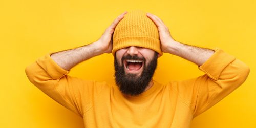 Laughing bearded hipster in sweater covering eyes with hat standing on yellow.